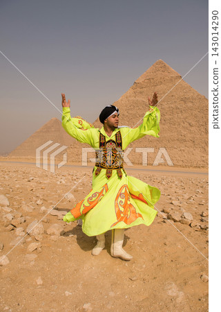 Young Egyptian tanoura folk dancer in vivid neon yellow robe spins arms raised before the iconic Great Pyramids of Giza desert plateau ideal for Egypt Young Egyptian tanoura folk dancer in vivid neon yellow robe spins arms raised before the iconic Great Pyramids of Giza desert plateau ideal for Egypt 143014290
