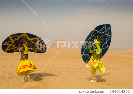 Two folk dancers in vibrant embroidered yellow costumes simultaneously raise giant geometric painted discs before the iconic Great Pyramids of Giza on Two folk dancers in vibrant embroidered yellow costumes simultaneously raise giant geometric painted discs before the iconic Great Pyramids of Giza on 143014291