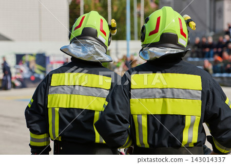 Two Firefighters in High-Visibility Helmets and Reflective Jackets Standing at Emergency Scene 143014337