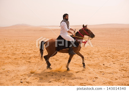 Authentic Arab horseman in traditional keffiyeh scarf riding bay horse at full gallop across a vast open desert landscape ideal for MENA equestrian Authentic Arab horseman in traditional keffiyeh scarf riding bay horse at full gallop across a vast open desert landscape ideal for MENA equestrian 143014340