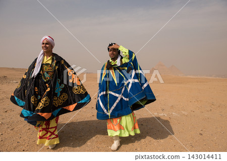 Two Egyptian tanoura folk performers wearing vivid ornate capes and turbans pose candidly before the iconic Great Pyramids of Giza on the desert Two Egyptian tanoura folk performers wearing vivid ornate capes and turbans pose candidly before the iconic Great Pyramids of Giza on the desert 143014411