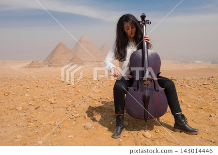 Soulful female cellist absorbed in performance seated on desert sand before the iconic Great Pyramids of Giza Egypt ideal for music arts and cultural Soulful female cellist absorbed in performance seated on desert sand before the iconic Great Pyramids of Giza Egypt ideal for music arts and cultural 143014446