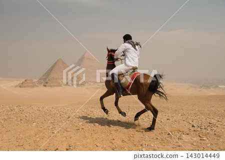 Arab horseman in white shirt and keffiyeh rides rearing arabian horse on sandy Giza plateau with three iconic ancient pyramids in hazy background Arab horseman in white shirt and keffiyeh rides rearing arabian horse on sandy Giza plateau with three iconic ancient pyramids in hazy background 143014449