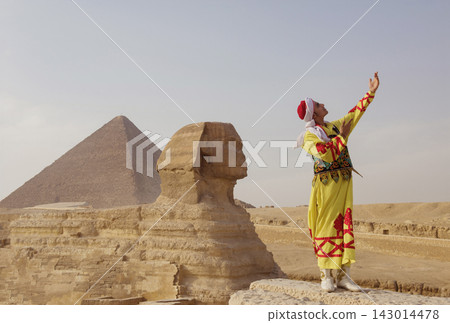 Joyful performer wearing vivid yellow red traditional Egyptian folk costume raises single arm skyward before the Great Sphinx and towering Khufu Joyful performer wearing vivid yellow red traditional Egyptian folk costume raises single arm skyward before the Great Sphinx and towering Khufu 143014478