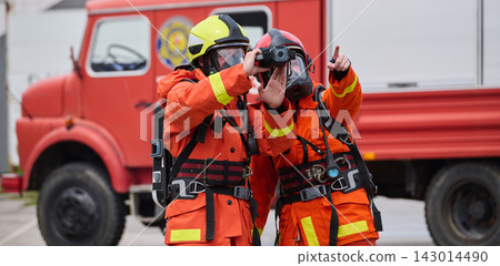 Two Firefighters in Orange Protective Gear Using Thermal Camera Beside Red Fire Truck During Emergency Response Two Firefighters in Orange Protective Gear Using Thermal Camera Beside Red Fire Truck During Emergency Response 143014490