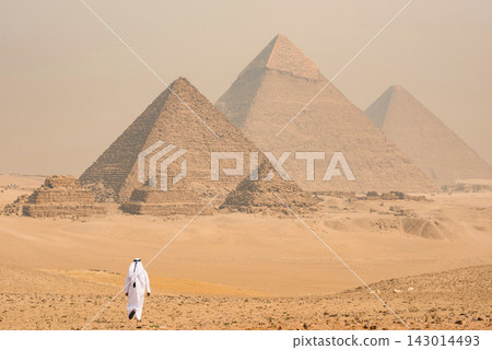 Solitary figure in traditional white thobe and agal headwear walking toward the three iconic Giza pyramids across sandy desert under hazy diffused sky Solitary figure in traditional white thobe and agal headwear walking toward the three iconic Giza pyramids across sandy desert under hazy diffused sky 143014493