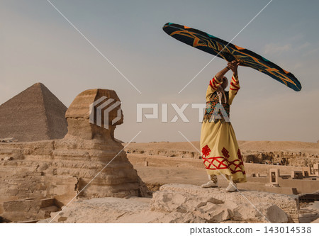 Traditional costumed performer dramatically raising ornate embroidered fabric before the Great Sphinx and Pyramid of Giza on the sun bleached plateau Traditional costumed performer dramatically raising ornate embroidered fabric before the Great Sphinx and Pyramid of Giza on the sun bleached plateau 143014538