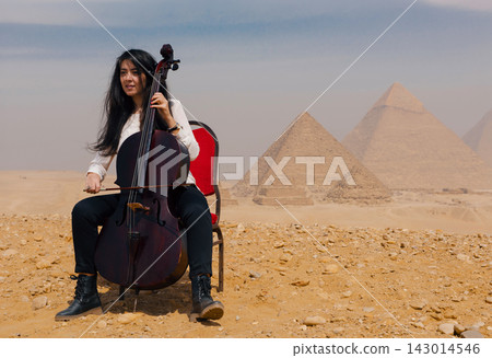 Young Middle Eastern woman playing cello seated on red chair before Great Pyramids of Giza on sandy desert plateau with overcast sky and dramatic Young Middle Eastern woman playing cello seated on red chair before Great Pyramids of Giza on sandy desert plateau with overcast sky and dramatic 143014546