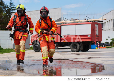 Firefighters in Full Gear Running Toward Emergency With Fire Truck and Water Splash During Training Drill Firefighters in Full Gear Running Toward Emergency With Fire Truck and Water Splash During Training Drill 143014550