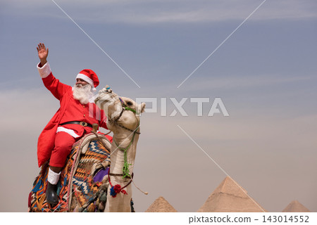 Joyful Santa Claus in full red costume waving triumphantly while riding an ornately decorated camel near the Great Pyramids of Giza under open sky Joyful Santa Claus in full red costume waving triumphantly while riding an ornately decorated camel near the Great Pyramids of Giza under open sky 143014552