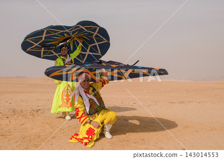 Two smiling kite festival performers in vibrant embroidered folk costumes hold massive geometric decorative kites aloft in open sandy desert ideal for Two smiling kite festival performers in vibrant embroidered folk costumes hold massive geometric decorative kites aloft in open sandy desert ideal for 143014553