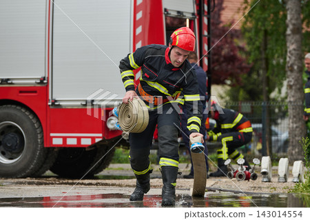 Firefighter Rolling Hose Near Fire Truck During Emergency Response Training With Team And Equipment Firefighter Rolling Hose Near Fire Truck During Emergency Response Training With Team And Equipment 143014554