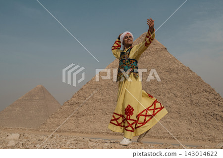 Smiling Arab male folk dancer in vibrant yellow embroidered robe and turban caught mid step with robe swirling before the iconic Great Pyramid of Giza 143014622