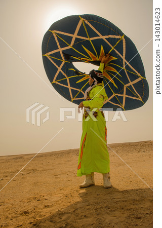 Colorful folk performer in ornate embroidered robe raises a massive geometric teal disc high overhead on sunlit Sahara desert sands ideal for cultural 143014623