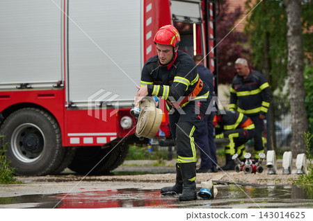 Firefighter Rolling Up Hose Beside Red Fire Truck During Emergency Rescue Operation Firefighter Rolling Up Hose Beside Red Fire Truck During Emergency Rescue Operation 143014625