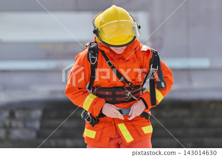 Firefighter In Orange Uniform And Yellow Helmet Fastening Safety Harness During Training Exercise Firefighter In Orange Uniform And Yellow Helmet Fastening Safety Harness During Training Exercise 143014630