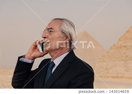 Composed silver haired senior executive in dark suit speaking on cell phone outdoors with iconic Great Pyramids of Giza softly blurred behind him Composed silver haired senior executive in dark suit speaking on cell phone outdoors with iconic Great Pyramids of Giza softly blurred behind him 143014659