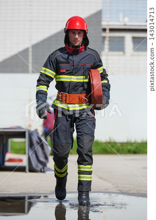 Firefighter In Protective Gear Walking With Hose And Red Helmet At Training Station Firefighter In Protective Gear Walking With Hose And Red Helmet At Training Station 143014713