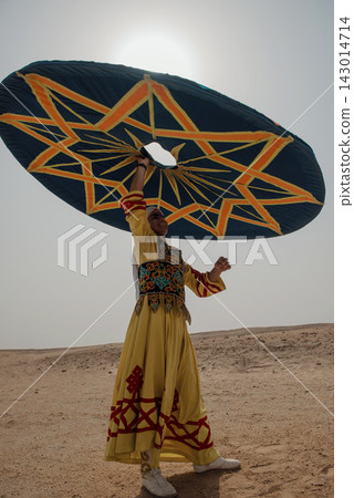 Egyptian tanoura dancer in yellow embroidered robe raises enormous teal geometric spinning disc overhead in open sandy desert under blazing midday sun Egyptian tanoura dancer in yellow embroidered robe raises enormous teal geometric spinning disc overhead in open sandy desert under blazing midday sun 143014714