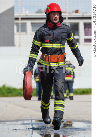 Firefighter Running With Hose Reel Wearing Helmet And Protective Gear During Emergency Drill Firefighter Running With Hose Reel Wearing Helmet And Protective Gear During Emergency Drill 143014730