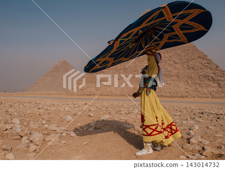 Cultural performer in vibrant embroidered yellow and red folk costume carrying large decorative prop overhead in silhouette walk before iconic Giza Cultural performer in vibrant embroidered yellow and red folk costume carrying large decorative prop overhead in silhouette walk before iconic Giza 143014732