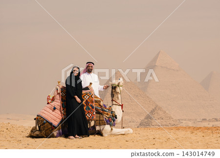 Arab couple in thobe abaya and keffiyeh posing happily with a decorated camel at the iconic Great Pyramids of Giza Egypt ideal for Gulf travel and Arab couple in thobe abaya and keffiyeh posing happily with a decorated camel at the iconic Great Pyramids of Giza Egypt ideal for Gulf travel and 143014749
