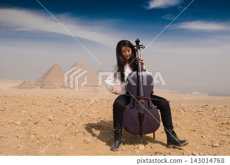 Focused young woman cellist actively bowing a cello while seated on the sandy Giza plateau with the iconic Great Pyramids of Giza dissolving into Focused young woman cellist actively bowing a cello while seated on the sandy Giza plateau with the iconic Great Pyramids of Giza dissolving into 143014768