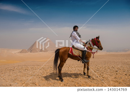 Commanding Arab horseman in white shirt and keffiyeh rides brown stallion on the Giza Plateau ancient pyramids rising from sandy desert haze ideal for Commanding Arab horseman in white shirt and keffiyeh rides brown stallion on the Giza Plateau ancient pyramids rising from sandy desert haze ideal for 143014769