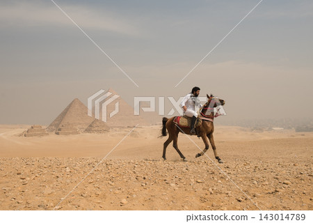 Confident Arab man on Arabian horse galloping across the Giza desert plateau with all three Great Pyramids in the background under a hazy overcast sky 143014789