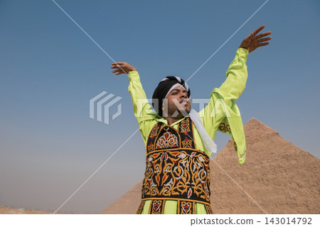 Egyptian cultural performer in neon yellow embroidered vest and turban raises arms in triumph before the Great Pyramid of Giza under a vivid blue sky 143014792