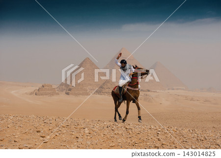 Exhilarated Arab horseman on galloping brown stallion raises arm before sweeping panorama of all three Giza pyramids on rocky sandy desert plateau Exhilarated Arab horseman on galloping brown stallion raises arm before sweeping panorama of all three Giza pyramids on rocky sandy desert plateau 143014825