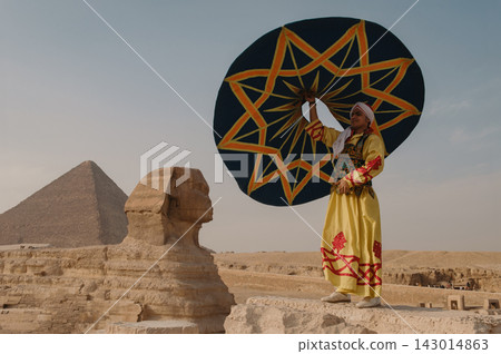 Proud Egyptian folk performer in ornate yellow robe and turban lifts a bold teal star patterned disc aloft beside the Great Sphinx of Giza with Proud Egyptian folk performer in ornate yellow robe and turban lifts a bold teal star patterned disc aloft beside the Great Sphinx of Giza with 143014863