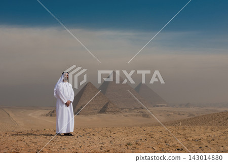 Proud Emirati man wearing white kandura and keffiyeh stands smiling before all three Giza Pyramids in Egypt with atmospheric desert haze and open blue Proud Emirati man wearing white kandura and keffiyeh stands smiling before all three Giza Pyramids in Egypt with atmospheric desert haze and open blue 143014880