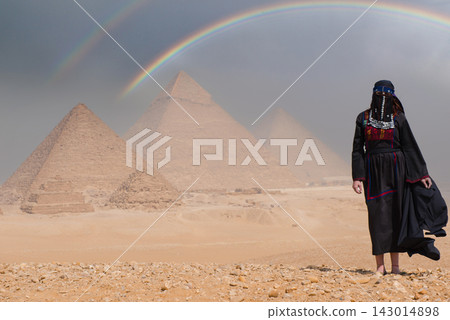 Traditionally veiled Egyptian woman in ornate folk costume stands on sandy Giza plateau gazing at the ancient pyramids beneath a breathtaking double Traditionally veiled Egyptian woman in ornate folk costume stands on sandy Giza plateau gazing at the ancient pyramids beneath a breathtaking double 143014898