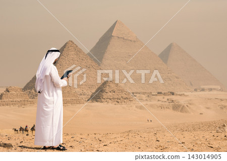 Rear view of Arab professional in white traditional kandura holding digital tablet while standing alone on sandy Giza plateau before three iconic Rear view of Arab professional in white traditional kandura holding digital tablet while standing alone on sandy Giza plateau before three iconic 143014905