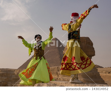 Two Egyptian folk dancers in ornate vivid costumes perform with arms outstretched before the Great Sphinx and Giza pyramid in a joyful cultural Two Egyptian folk dancers in ornate vivid costumes perform with arms outstretched before the Great Sphinx and Giza pyramid in a joyful cultural 143014952
