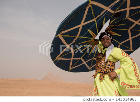Smiling Egyptian tanoura dancer in vivid lime embroidered robe holds giant decorative umbrella on the Giza desert plateau with pyramids in background Smiling Egyptian tanoura dancer in vivid lime embroidered robe holds giant decorative umbrella on the Giza desert plateau with pyramids in background 143014963