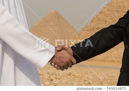 Close up of Arab man in white thobe and Western executive in dark suit shaking hands before ancient Giza pyramids representing cross cultural Close up of Arab man in white thobe and Western executive in dark suit shaking hands before ancient Giza pyramids representing cross cultural 143014973