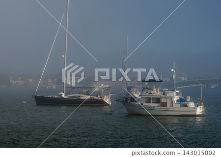 A dark sailboat and white motorboat rest on the water near Saint Augustine waterfront homes. Morning fog covers the shoreline and creates a quiet coastal scene. A dark sailboat and white motorboat rest on the water near Saint Augustine waterfront homes. Morning fog covers the shoreline and creates a quiet coastal scene. 143015002