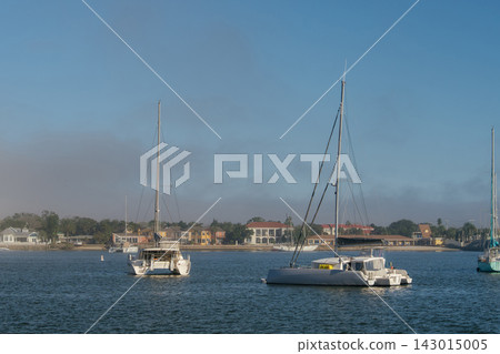 Two large catamarans float on blue water near the Saint Augustine shoreline. Colorful waterfront homes and low morning fog appear behind the anchored boats. Two large catamarans float on blue water near the Saint Augustine shoreline. Colorful waterfront homes and low morning fog appear behind the anchored boats. 143015005