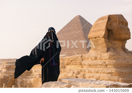 Dramatic portrait of a woman in traditional black niqab and flowing abaya with coin jewelry standing before Great Sphinx and Giza pyramid on sandy Dramatic portrait of a woman in traditional black niqab and flowing abaya with coin jewelry standing before Great Sphinx and Giza pyramid on sandy 143015009