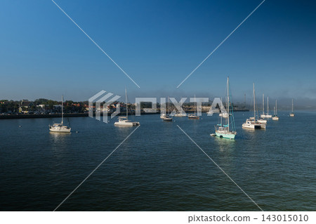 Numerous sailboats and catamarans float across the broad blue harbor in Saint Augustine, Florida. The historic waterfront and low coastal haze appear along the distant shoreline. Numerous sailboats and catamarans float across the broad blue harbor in Saint Augustine, Florida. The historic waterfront and low coastal haze appear along the distant shoreline. 143015010