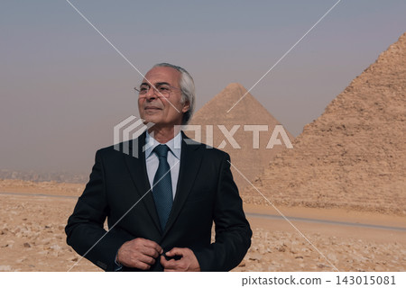Silver haired senior executive in dark navy suit and glasses stands confidently before the Great Pyramids of Giza gazing upward with composed Silver haired senior executive in dark navy suit and glasses stands confidently before the Great Pyramids of Giza gazing upward with composed 143015081