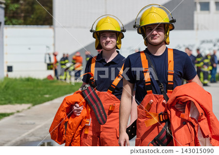 Two Young Firefighters in Yellow Helmets and Orange Protective Gear During Training Exercise Outdoors Two Young Firefighters in Yellow Helmets and Orange Protective Gear During Training Exercise Outdoors 143015090