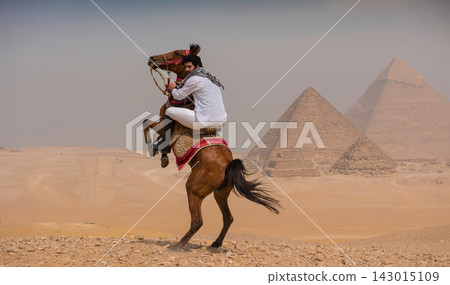 Dramatic Arab horseman in white shirt and keffiyeh rides a rearing horse on the sandy Giza plateau with iconic pyramids looming in hazy background Dramatic Arab horseman in white shirt and keffiyeh rides a rearing horse on the sandy Giza plateau with iconic pyramids looming in hazy background 143015109