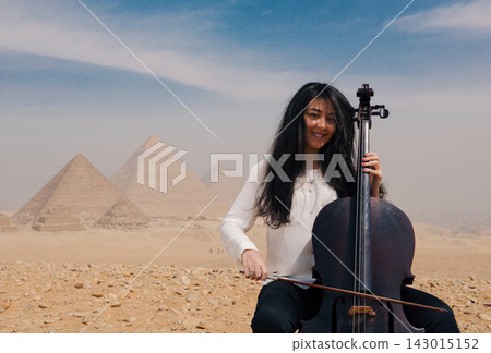 Young Middle Eastern woman cellist performing with joyful smile before the iconic Giza pyramids on a sunny desert plateau in Egypt ideal for arts and Young Middle Eastern woman cellist performing with joyful smile before the iconic Giza pyramids on a sunny desert plateau in Egypt ideal for arts and 143015152
