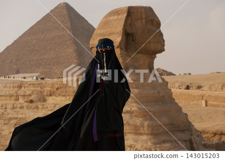 Veiled niqab woman in dramatic wind swept black abaya adorned with silver coin jewelry stands before the Great Sphinx of Giza with the Khafre pyramid Veiled niqab woman in dramatic wind swept black abaya adorned with silver coin jewelry stands before the Great Sphinx of Giza with the Khafre pyramid 143015203