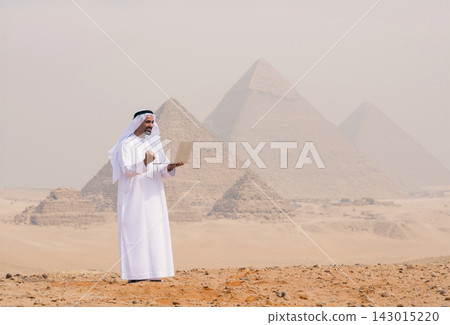 Ambitious Arab entrepreneur in traditional white kandura and ghutra using a laptop confidently outdoors on the sandy Giza plateau with the iconic Ambitious Arab entrepreneur in traditional white kandura and ghutra using a laptop confidently outdoors on the sandy Giza plateau with the iconic 143015220