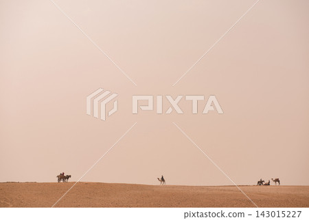 Camel riders and horse cart traverse a sweeping Egyptian desert plateau under enormous hazy pale sky ideal for MENA travel editorial and minimalist Camel riders and horse cart traverse a sweeping Egyptian desert plateau under enormous hazy pale sky ideal for MENA travel editorial and minimalist 143015227