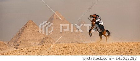 Fearless Arab horseman on a fully rearing Arabian horse in sharp focus before the iconic Giza pyramid complex across a vast golden sandy plateau in Fearless Arab horseman on a fully rearing Arabian horse in sharp focus before the iconic Giza pyramid complex across a vast golden sandy plateau in 143015288